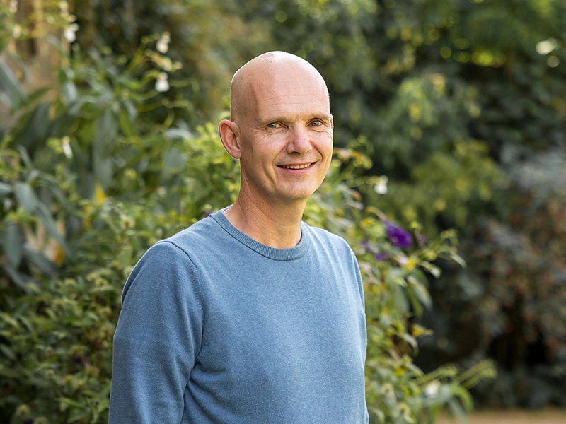 A man in a blue sweater poses for a portrait at a green garden.
