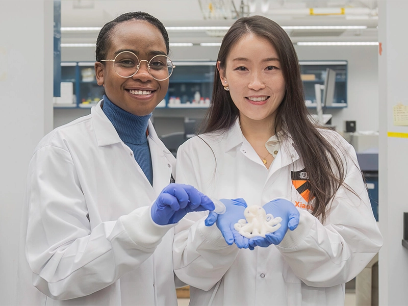 Two women in lab coats hold a hydrogel shaped like a cartoon octopus.