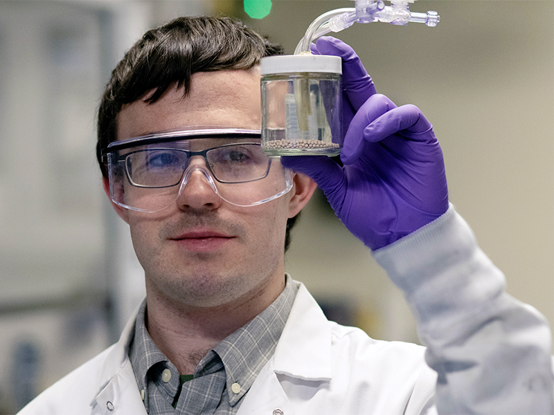 A man in a lab coat and goggles holds up a small jar containing pinkish pebble-like material.
