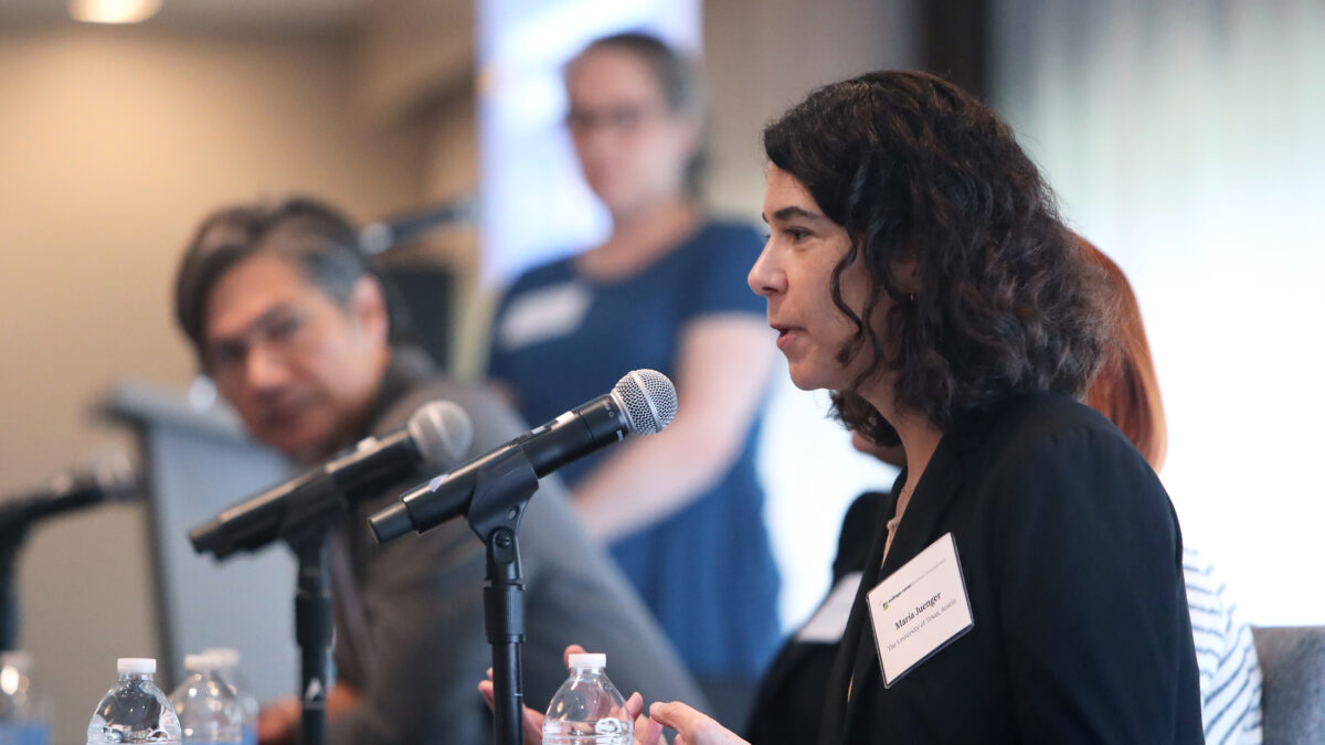 A woman seated at a microphoned panel talks as a man and a woman look on.