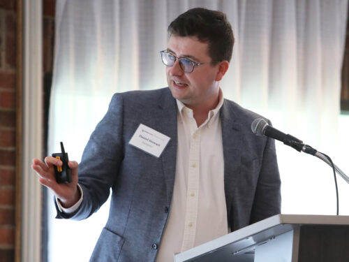 A man in a grey suit at a podium gestures to his right side.