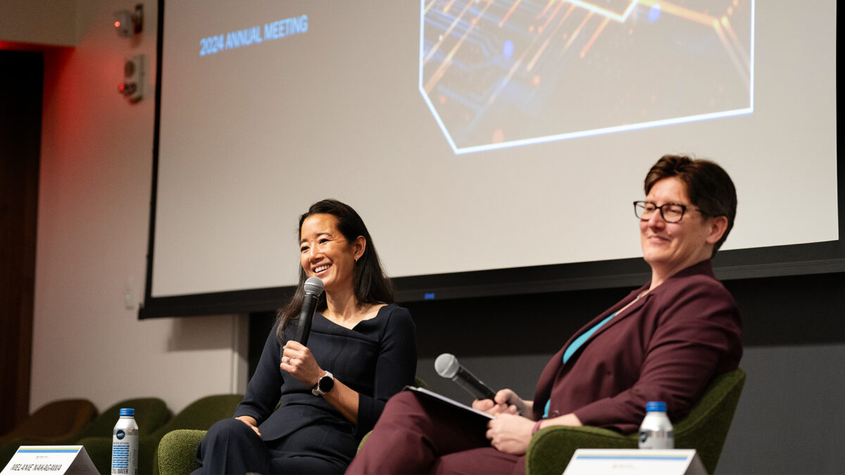 Two women seated on a stage. One speaks to the crowd using a microphone.