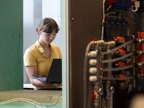 A woman in goggles and surrounded by machinery holds a laptop.