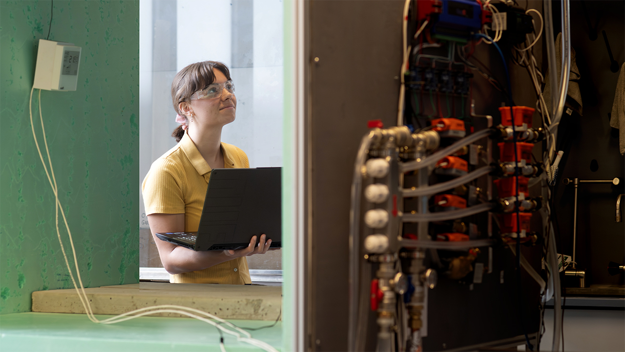 A woman in goggles and surrounded by machinery holds a laptop.