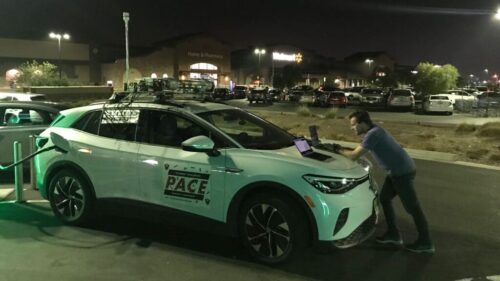 A man works on a laptop open on a car's hood in a parking lot