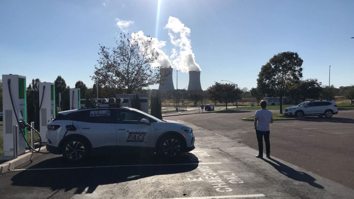 Man stands in front of an electric vehicle at a charging station with cooling towers in the distance