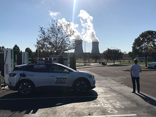 Man stands in front of an electric vehicle at a charging station with cooling towers in the distance