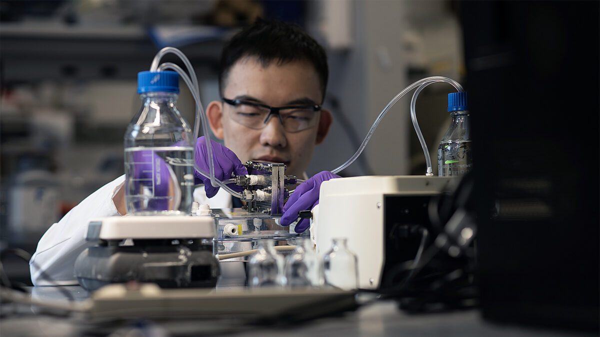 A man in a lab coat and safety glasses operates a machine in a laboratory involving bottles of clear liquid and tubing.