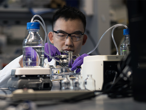 A man in a lab coat and safety glasses operates a machine in a laboratory involving bottles of clear liquid and tubing.