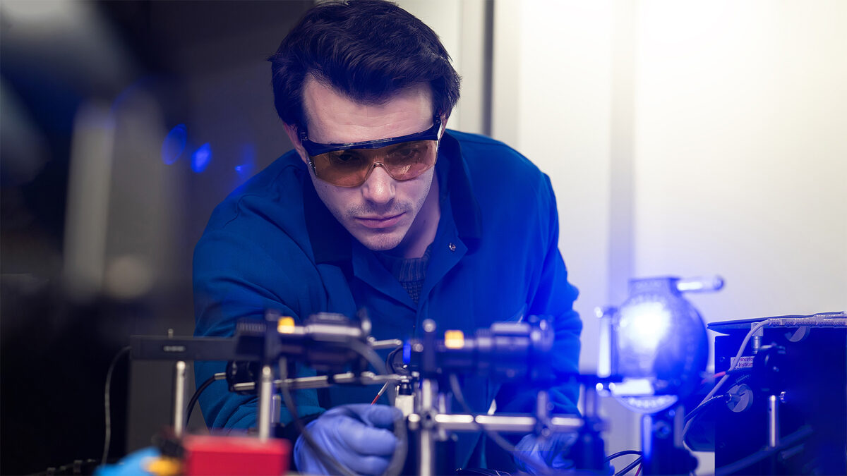 A researcher wearing safety goggles and gloves examines a complex laser setup in a lab. The scene is brightly lit, conveying focus and innovation.