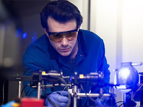 A researcher wearing safety goggles and gloves examines a complex laser setup in a lab. The scene is brightly lit, conveying focus and innovation.