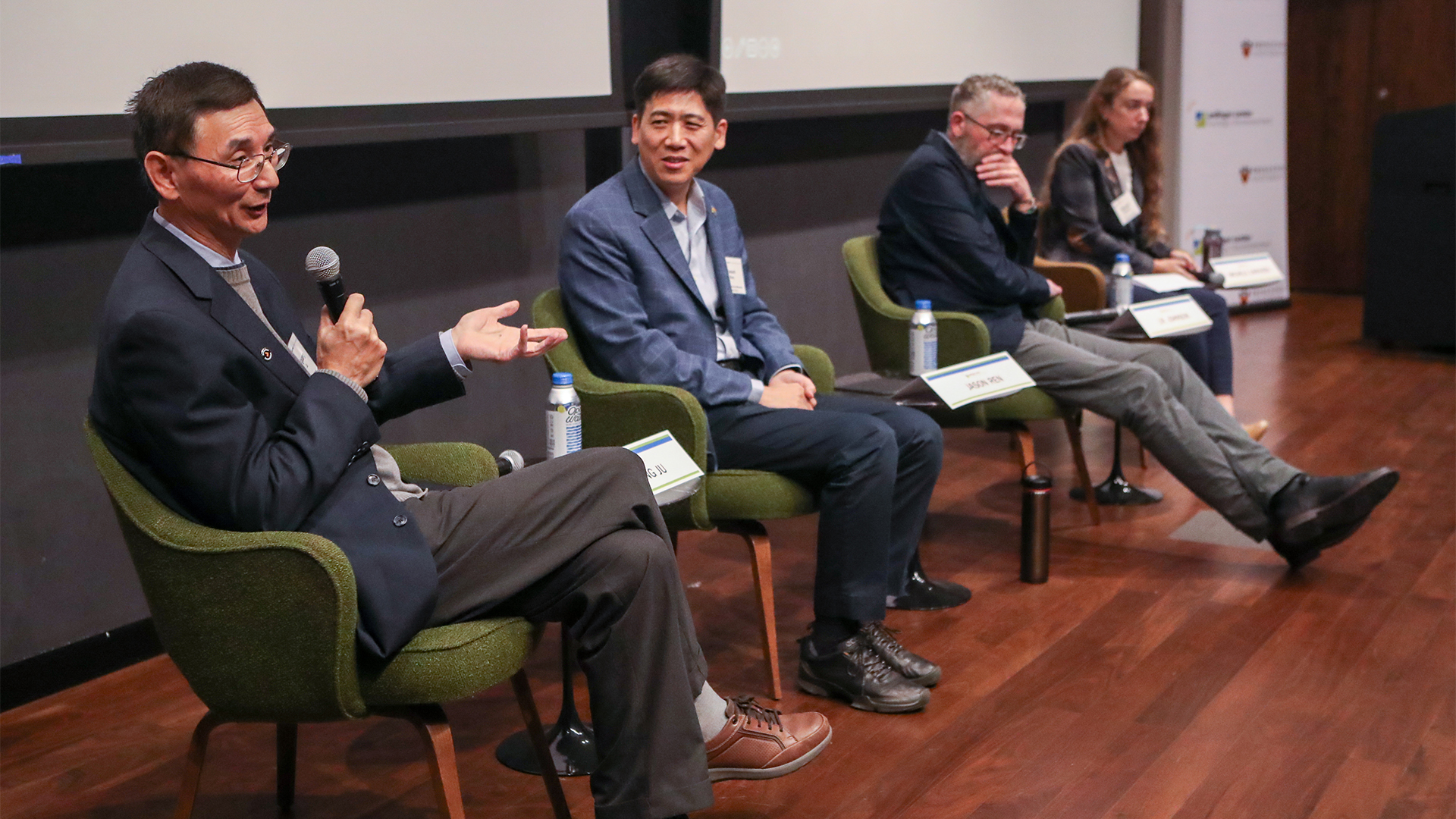 Four panelists sit on stage, each in green chairs, engaged in discussion. One man speaks into a microphone, while others listen attentively. The setting is formal.