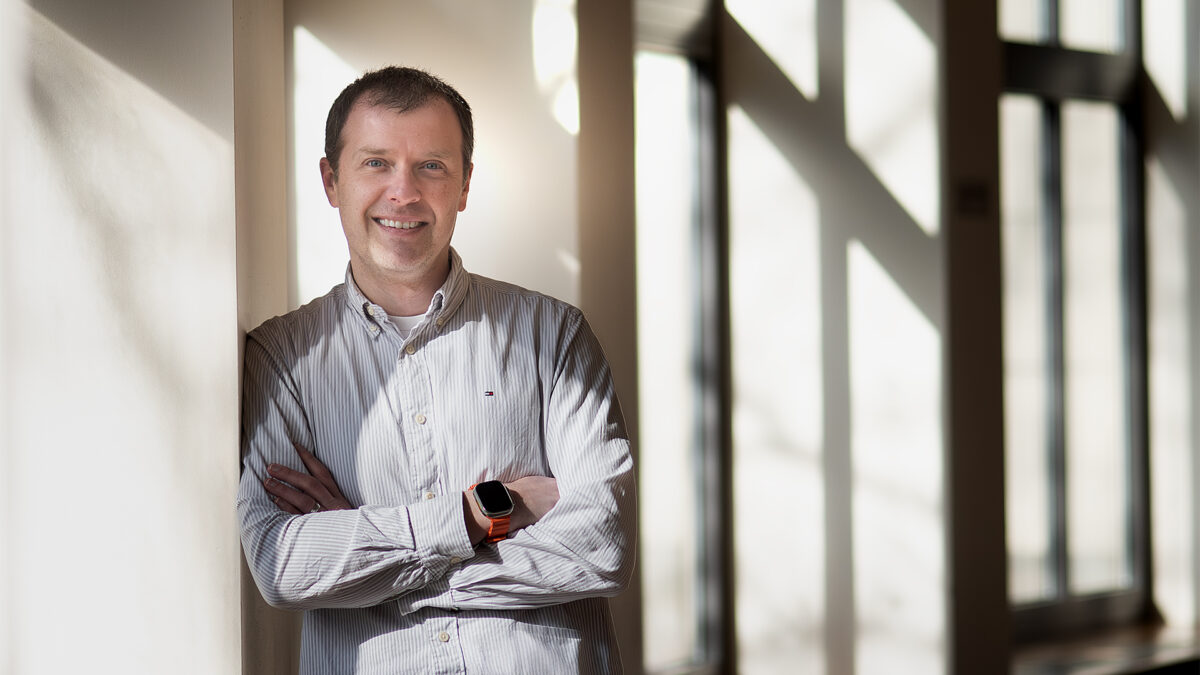A man stands with his arms crossed in a bright room with many windows.