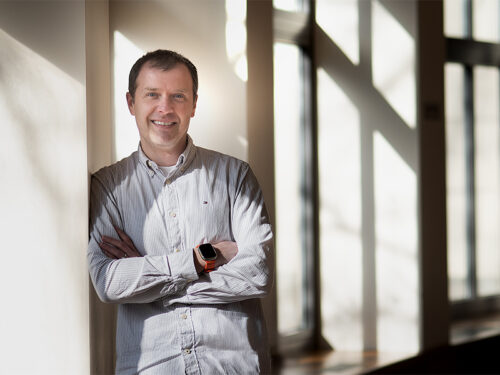 A man stands with his arms crossed in a bright room with many windows.