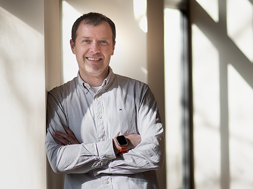 A man stands with his arms crossed in a bright room with many windows.