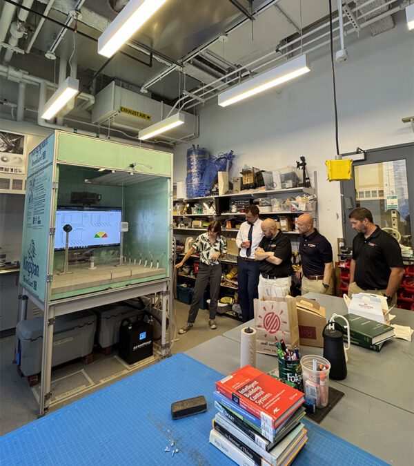 A group of people stand in a cluttered workshop, observing a machine displaying a graph on a screen. 