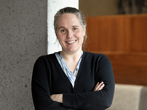 A woman stands with her arms crossed in a modern lobby setting.
