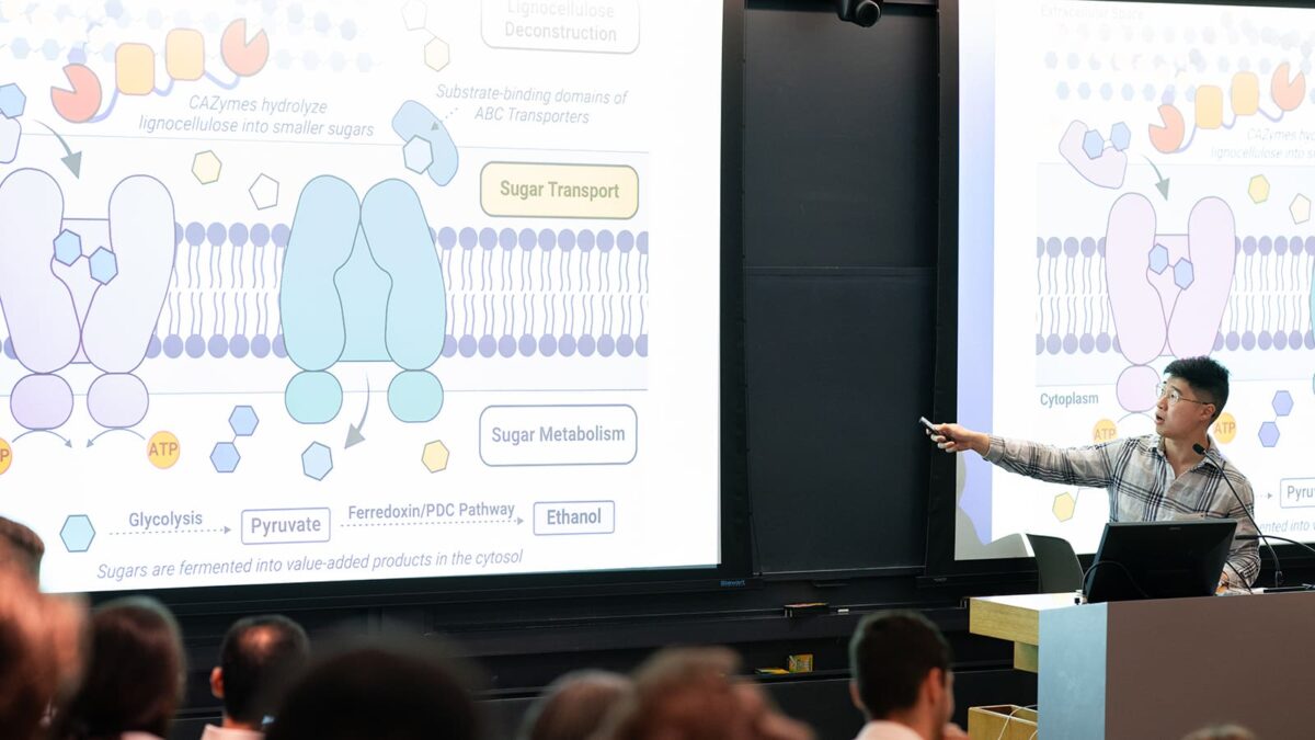 A person is giving a lecture, pointing at a projected slide with complex biochemical diagrams.