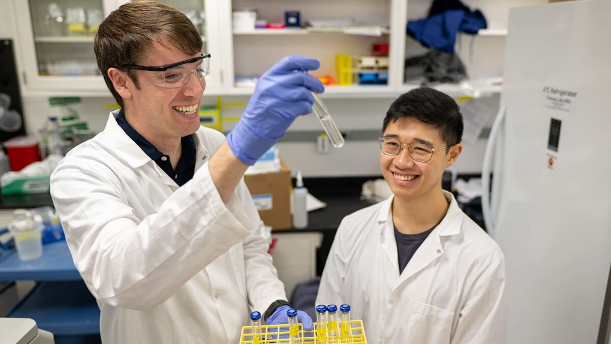 Two scientists in lab coats are in a laboratory. One, holding a test tube and wearing protective goggles, smiles at a colleague, who is also smiling.