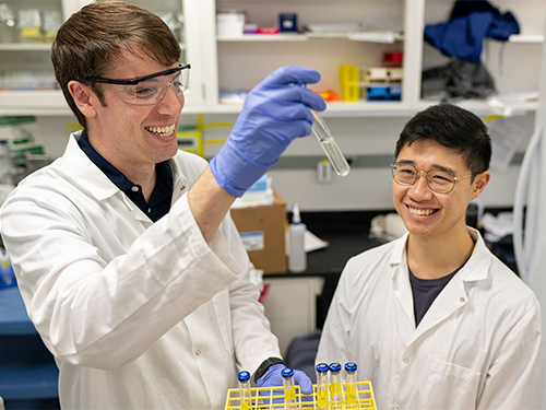 Two scientists in lab coats and goggles smile as one holds up a test tube.