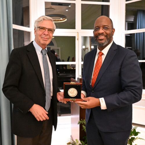 Two men in formal attire stand smiling indoors. One man holds an award plaque. 