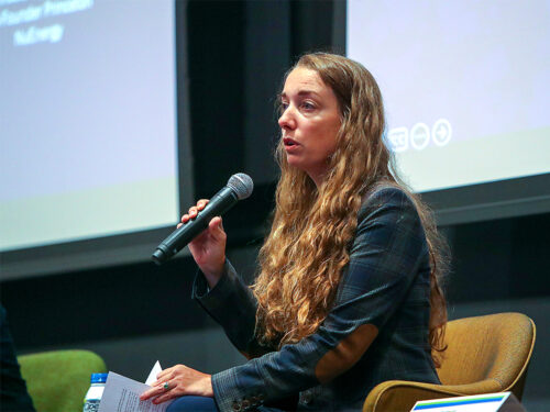 A woman with long, wavy hair speaks into a microphone during a panel discussion. She sits on a chair, holding notes.