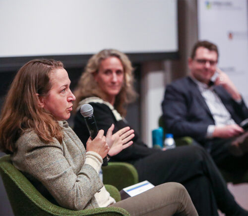 A woman speaks into a microphone during a panel discussion, seated in front of two listening colleagues.