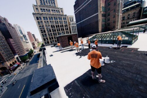 A group of people in orange shirts paint a rooftop white, surrounded by tall buildings.