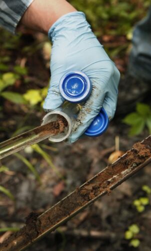 A gloved hand holds a test tube and a soil sample tool, collecting earthy soil. 