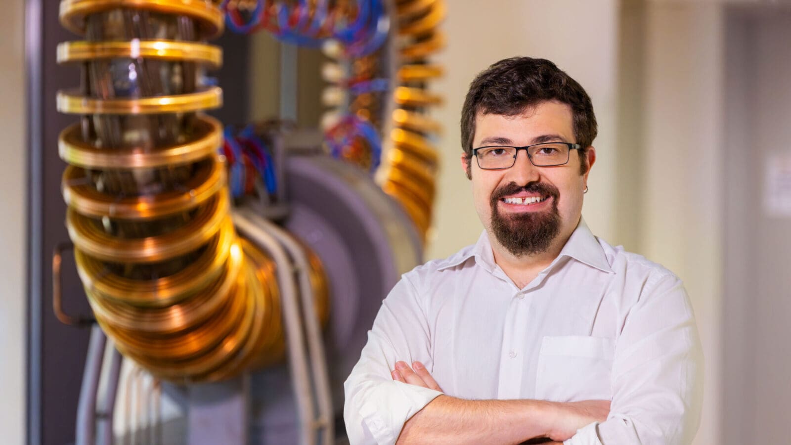 A bearded man wearing glasses stands before a large machine with coils