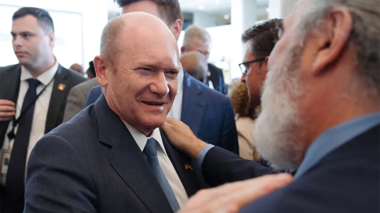 A man in a suit smiles warmly in a crowded event, receiving a friendly pat on the shoulder from another man. The atmosphere is friendly and engaging.