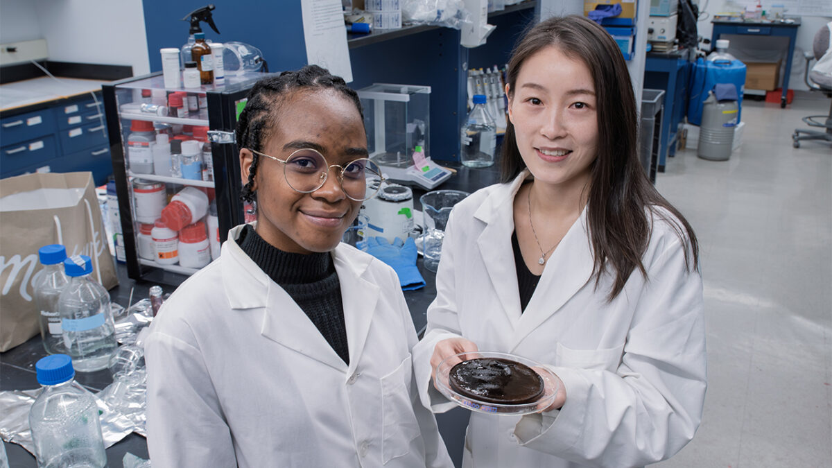 Two women in lab coats stand in a laboratory. One holds a petri dish with a brown substance, surrounded by scientific equipment and shelves.