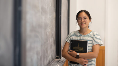 A young woman clutches notebooks next to a blackboard.