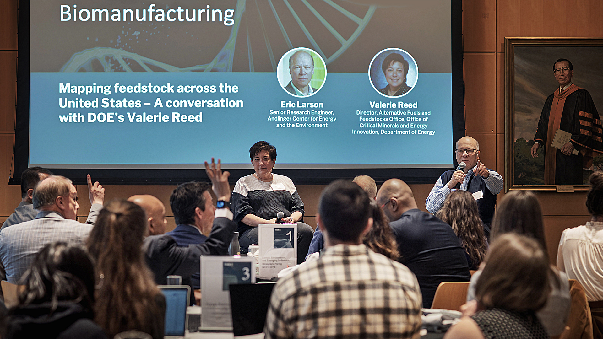 Audience attending a conference on biomanufacturing with speakers Eric Larson and Valerie Reed featured on a screen.