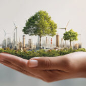 A hand holds a miniature cityscape with trees, wind turbines, and industrial buildings.