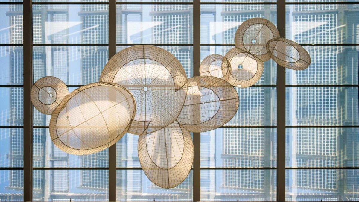 Hanging installation of large, woven, circular structures casting intricate shadows against a tall glass building.