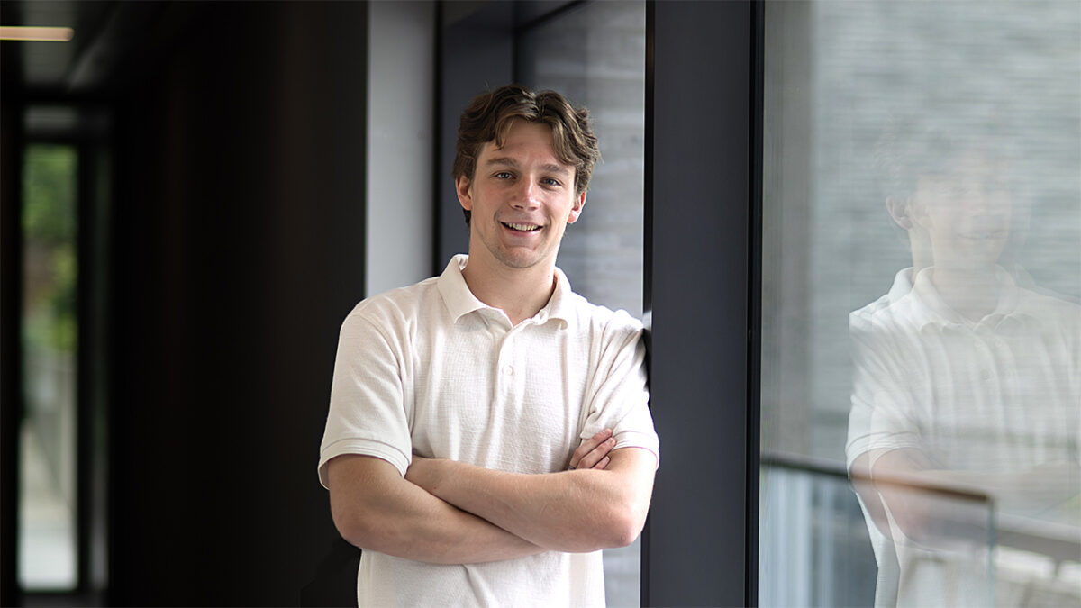 A young man stands by a large glass wall.