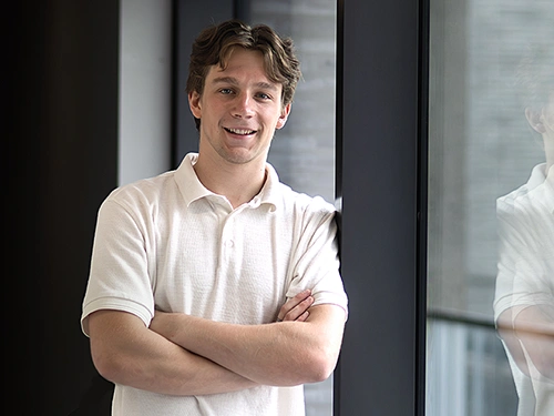 A young man stands by a large glass wall.