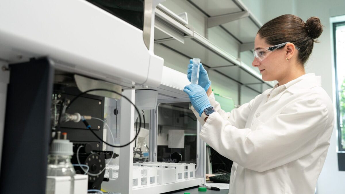 A young woman in a white lab coat examines a test tube near a fume hood.