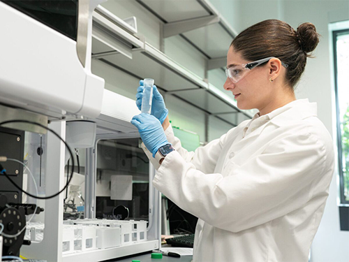 A young woman in a white lab coat examines a test tube near a fume hood.