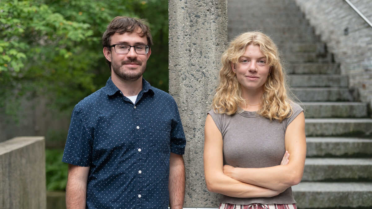 Two casually dressed adults pose outdoors beside a stone column and steps: a bearded man smiles gently, while a woman stands arms crossed.