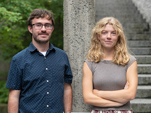 Two casually dressed adults pose outdoors beside a stone column and steps: a bearded man smiles gently, while a woman stands arms crossed.