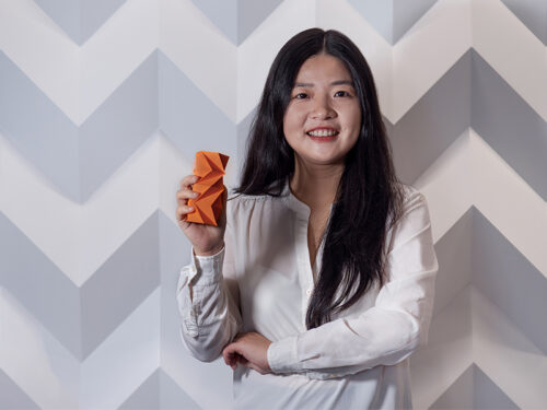 A woman holds an orange origami cylinder against a geometric wall.