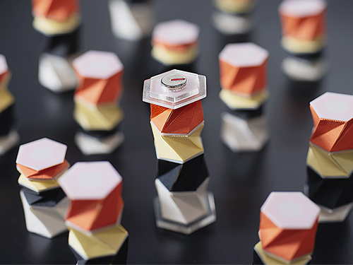 A close-up of orange, yellow, and white geometric origami pegs on a dark board, with one central piece topped by a small magnet.