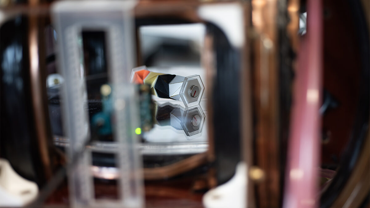 A compact silver device with two hexagonal components sits behind reflective glass in a small mechanical housing.