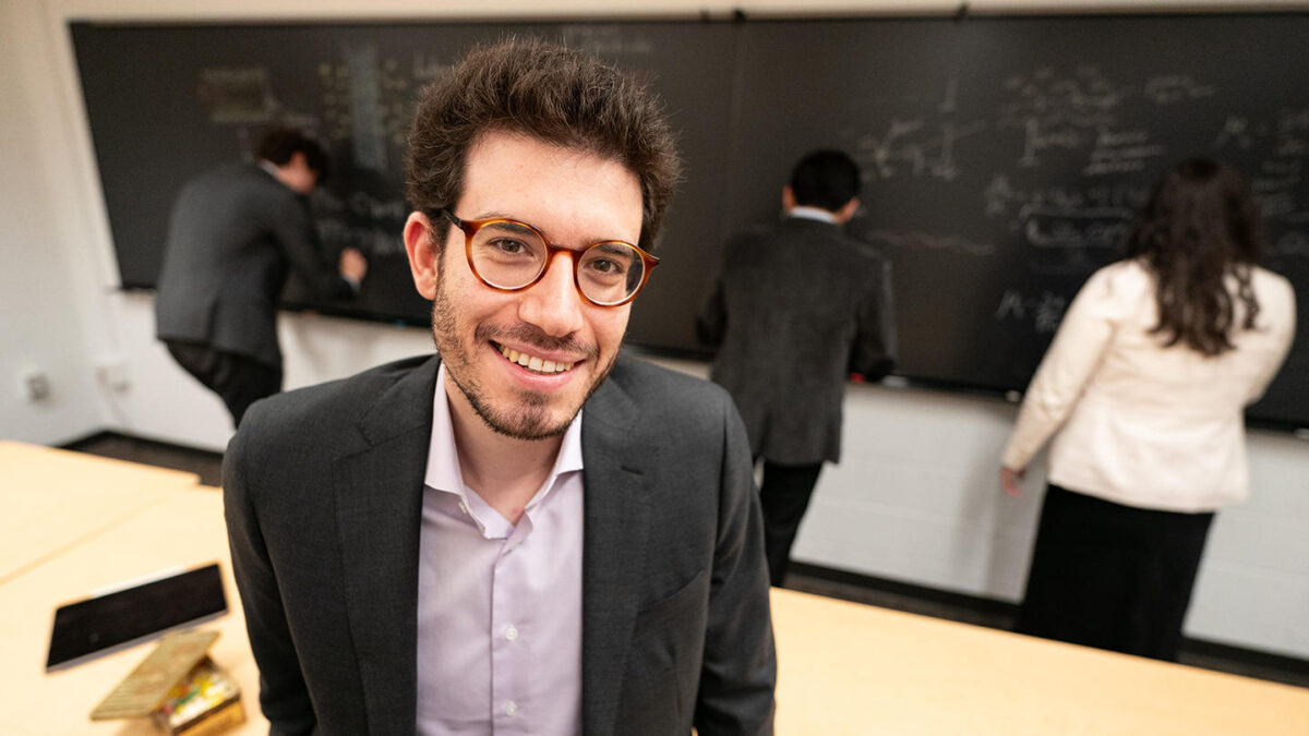 A man in glasses smiles at the camera as three individuals write on a chalkboard behind him.