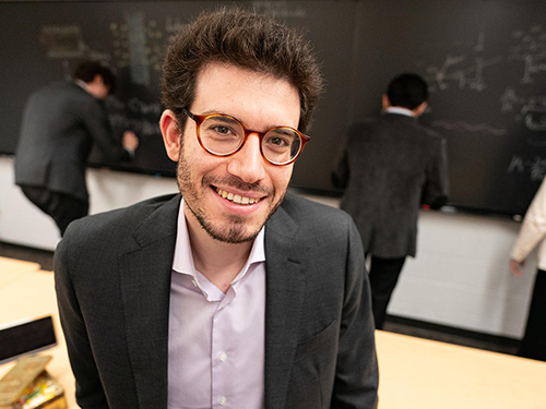 A man in glasses smiles at the camera as three individuals write on a chalkboard behind him.